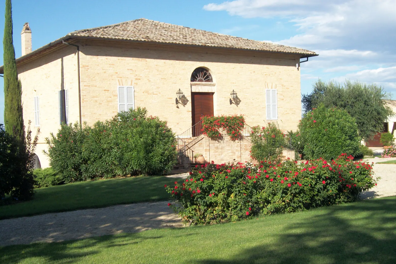 La facciata in mattoni della villa con la scalinata d'ingresso, gli oleandri e le rose rosse sul prato