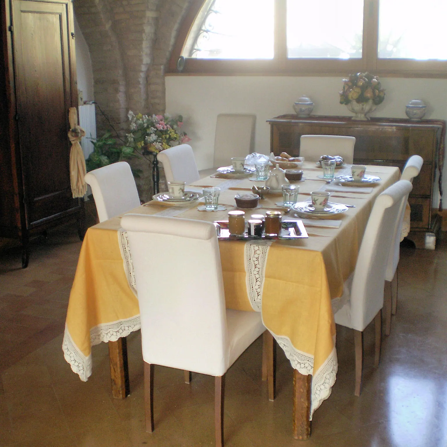 La tavola della colazione apparecchiata con tovaglia gialla, tazze, marmellate e dolci, davanti alla finestra ad arco